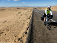 A bicycle tourist rides on the shoulder of a road with sand dunes on either side.
