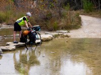 A bicyclist uses stepping stones to cross a stream while pushing her bicycle