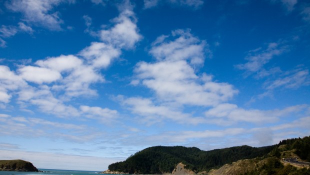 A view of deep blue sky with white fluffy clouds over a hill with a road winding down on the right side. The ocean is on the left with waves crashing ashore.