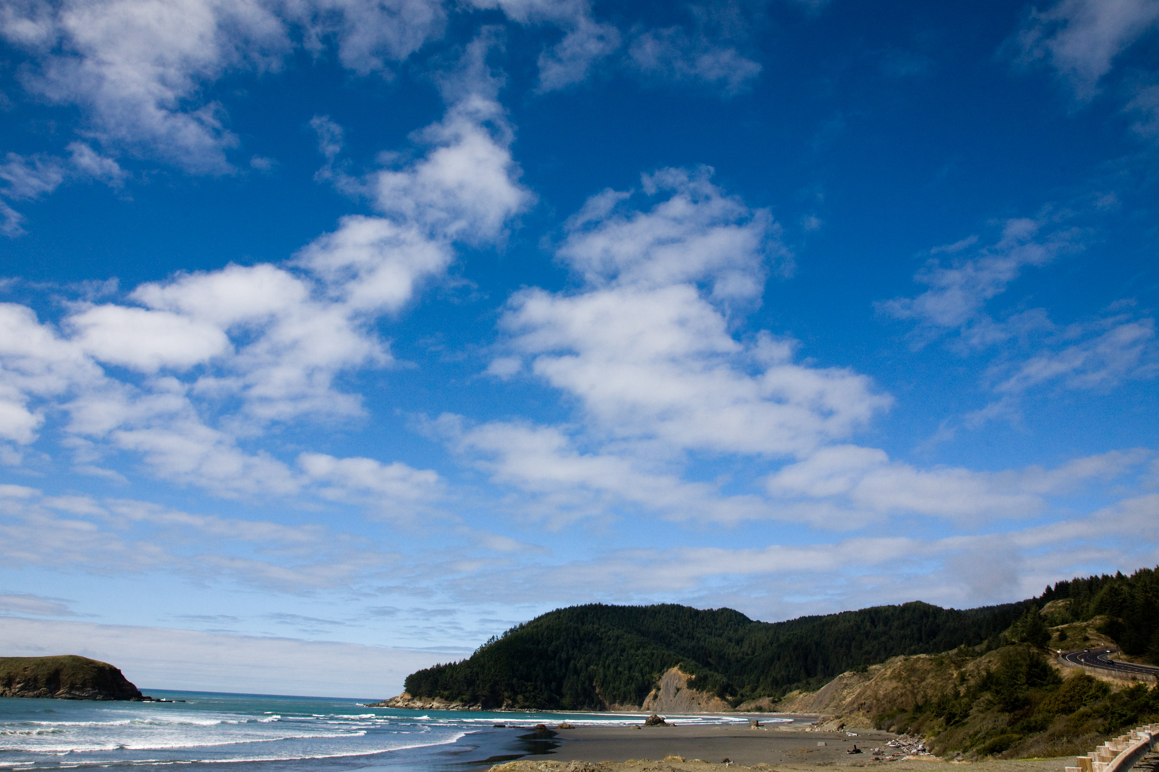 A view of deep blue sky with white fluffy clouds over a hill with a road winding down on the right side. The ocean is on the left with waves crashing ashore.