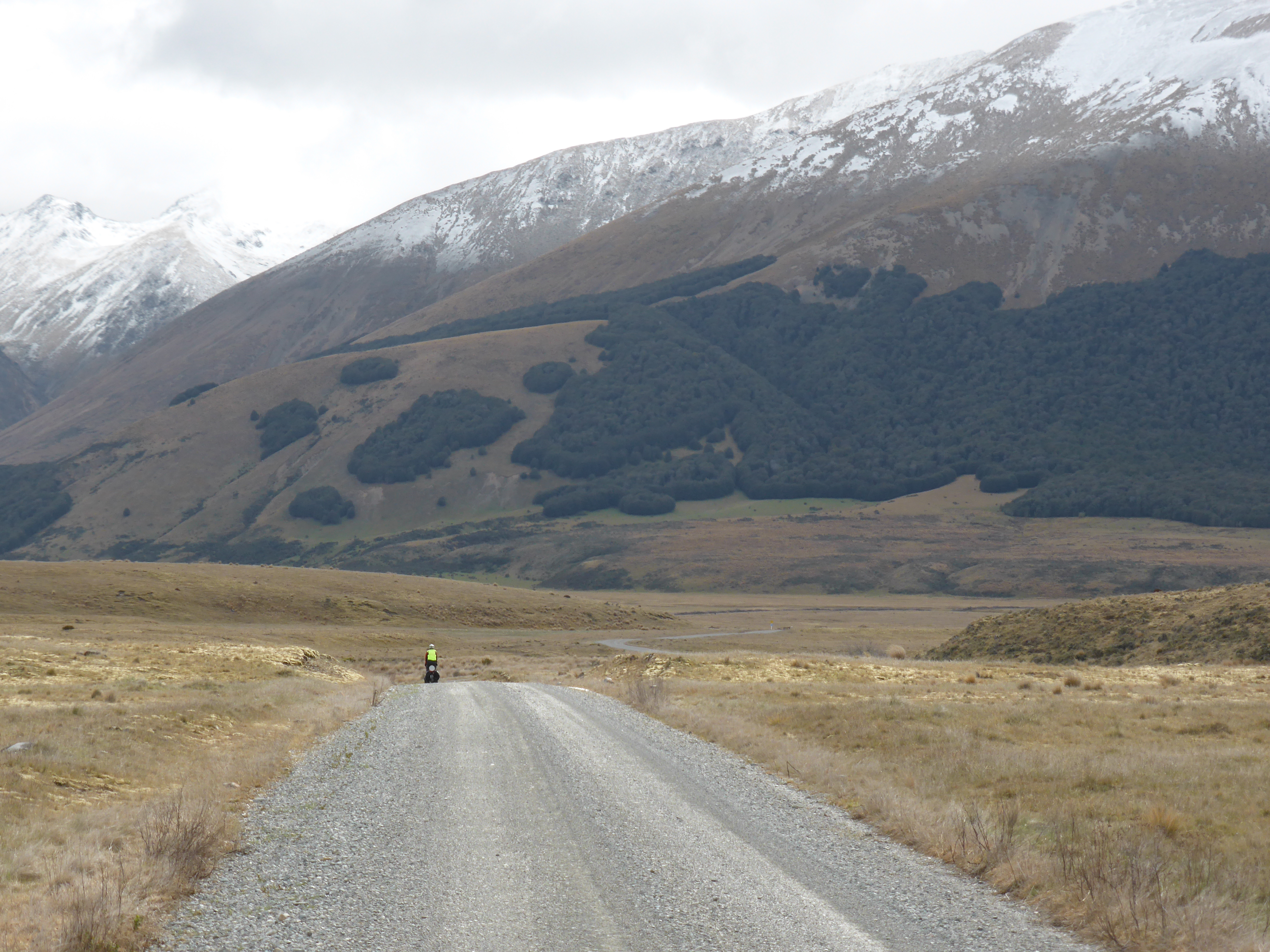 A cyclist rides a gravel road with snow capped mountains surrounding