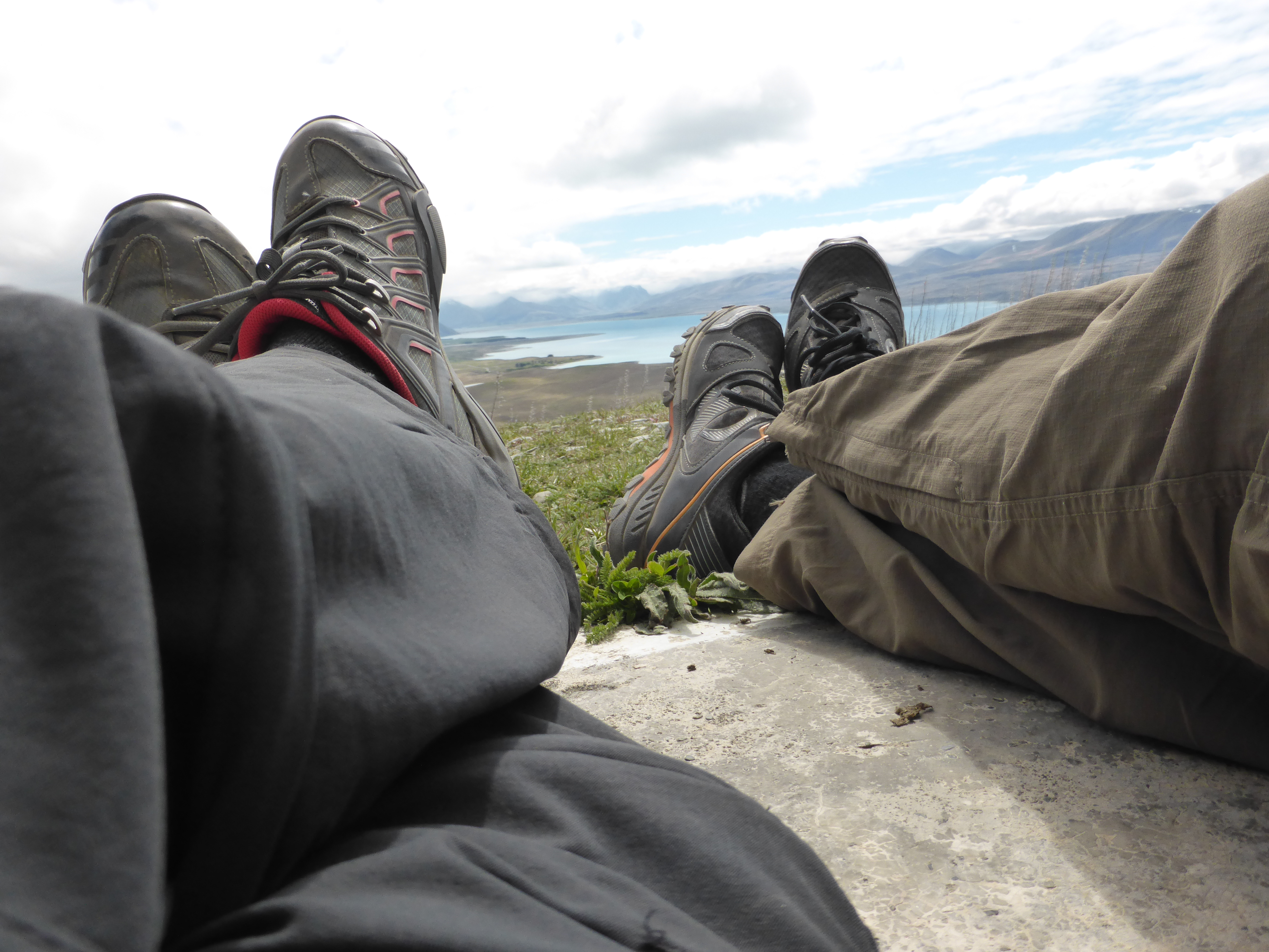 Lake Tekapo in the distance, two sets of legs and shoes in the foreground