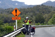 A bicyclist stands next to a road sign which reads "slumps"