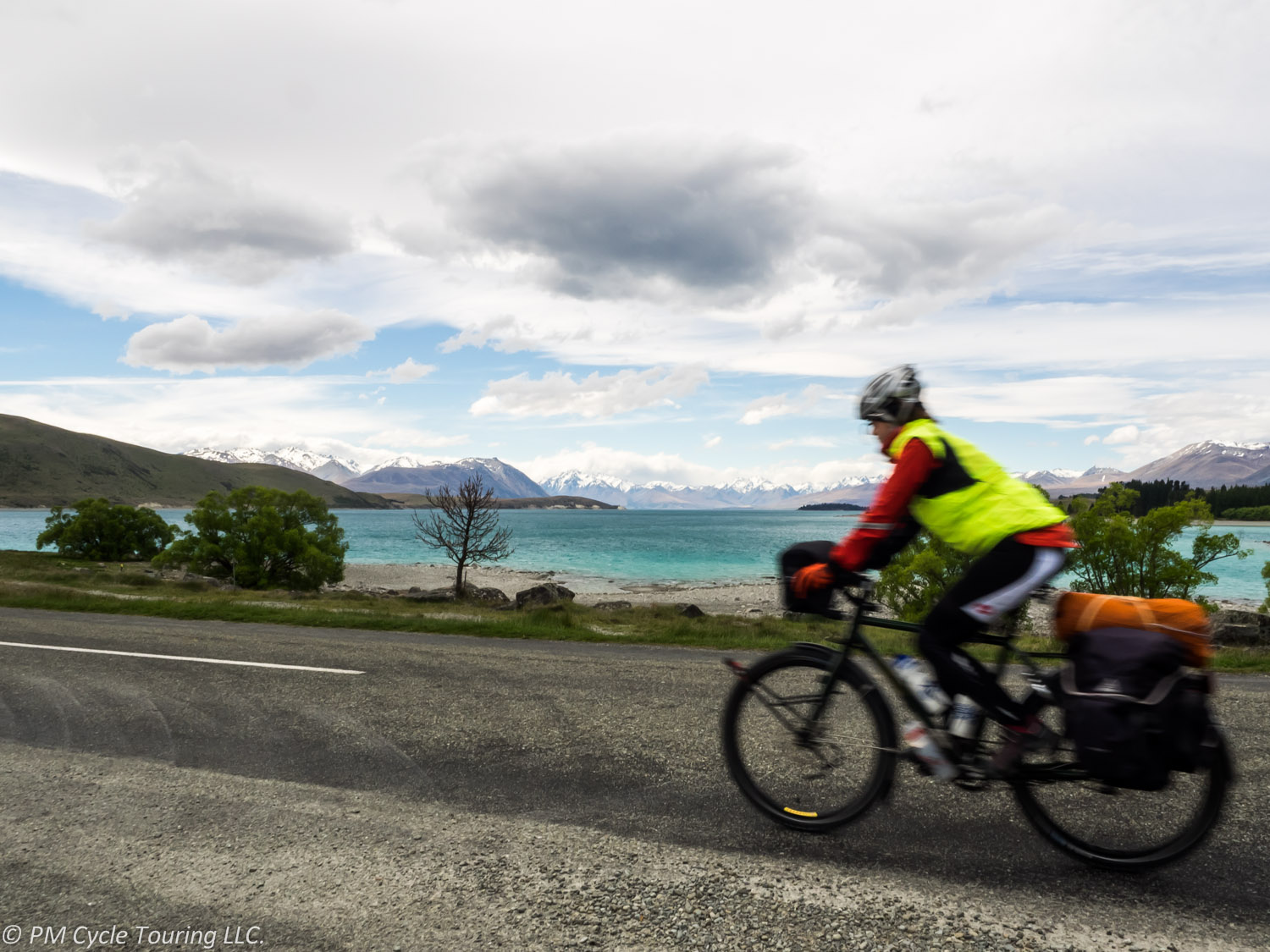 A woman on a fully loaded touring bicycle rides in front of Lake Tekapo, New Zealand